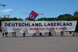 Eine Gruppe von Menschen mit einem Transparent, auf dem "Deutschland, Lagerland Migration ist kein Verbrechen" steht, und einer Flagge, die auf dem Boden steht, mit Straßenlaternen, Bäumen, Kränen, einem Gebäude und einem bewölkten Himmel im Hintergrund.