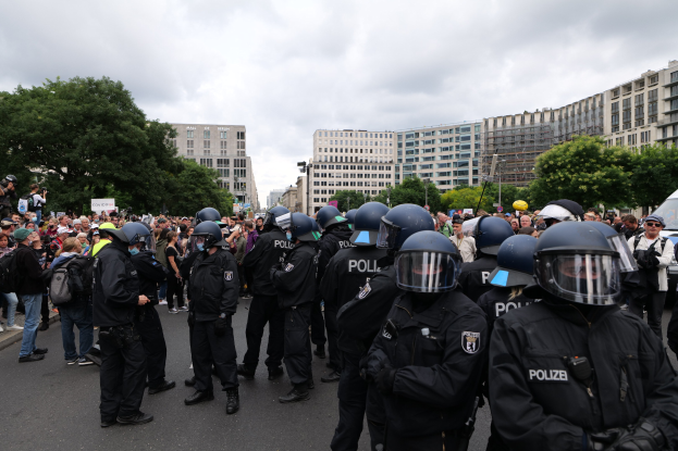 Große Gruppe von Polizeibeamten bei einer Demonstration in Berlin, Deutschland, mit einigen Personen, die Kameras halten, sowie Bäumen und Gebäuden im Hintergrund bei bewölktem Himmel.