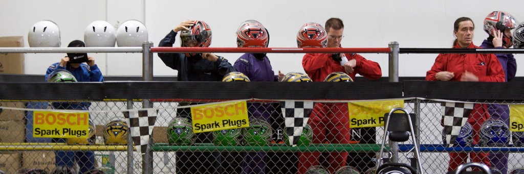 Ein Maschendrahtzaun mit Plakaten und Fahnen daran, einige Menschen im Hintergrund, einige tragen Helme, und Helme auf einem Ständer, mit einer Wand im Hintergrund.
