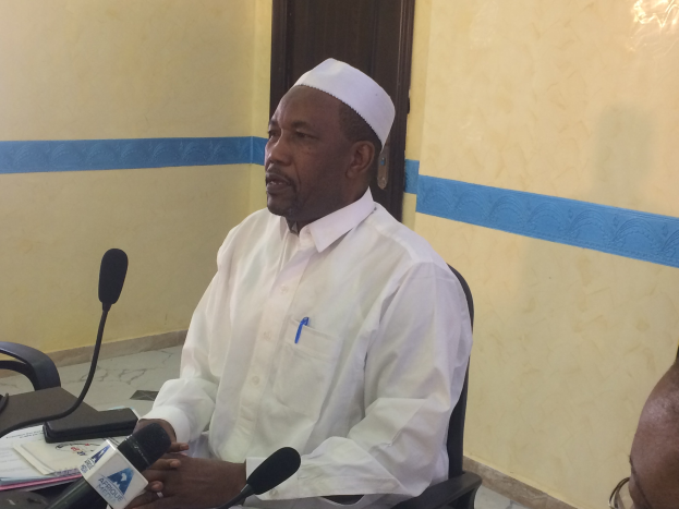 Ein Mann in einem weißen Kleid und Hut sitzt an einem Tisch mit Mikrofonen und spricht, während Papiere und Gegenstände auf dem Tisch zu sehen sind, mit einer anderen Person mit Brille zu seiner Rechten in einem Raum mit einer Tür im Hintergrund.