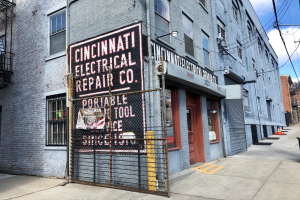 Außenansicht des Cincinnati Electrical Repair Co.-Gebäudes mit Fenstern, Türen, einem Schild, einem Metallzaun, einem Müllcontainer, Pfählen, Drähten, Bäumen und einem bewölkten Himmel.