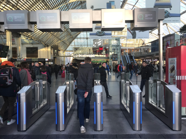 Eine Gruppe von Menschen geht durch einen belebten Bahnhof mit Metalldetektoren, Texttafeln, Säulen, Rolltreppen und Drehkreuzen unter einer sichtbaren Decke.