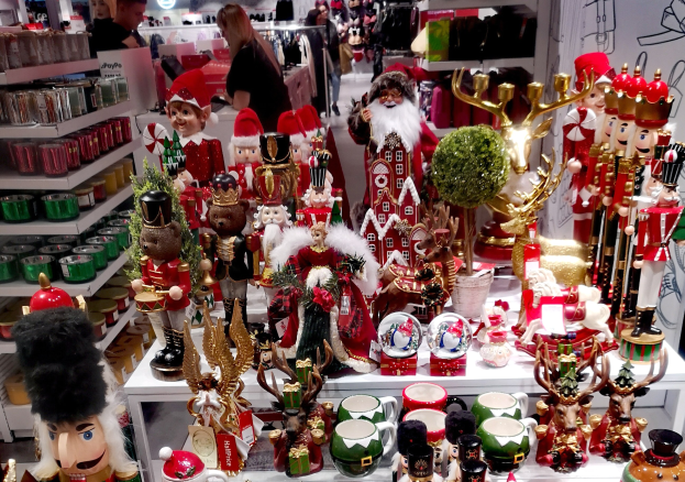 Ladeninneneinrichtung mit Weihnachtsdekorationen, einschließlich Spielzeug, Tassen und anderen Gegenständen auf Tischen und Regalen, mit Menschen, Lichtern und einer Wand im Hintergrund.