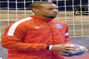 Ein Mann in einem blauen T-Shirt und Shorts steht vor einem Tor, hält einen Fussball und hat einen entschlossenen Gesichtsausdruck, seine kurzen dunklen Haare und konzentrierten Augen auf den Ball gerichtet.