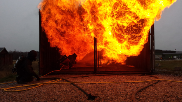 Feuerwehrleute löschen ein Gebäudebrand mit einem Schlauch auf dem Boden, Schuppen und Bäume im Hintergrund unter einem klaren blauen Himmel.