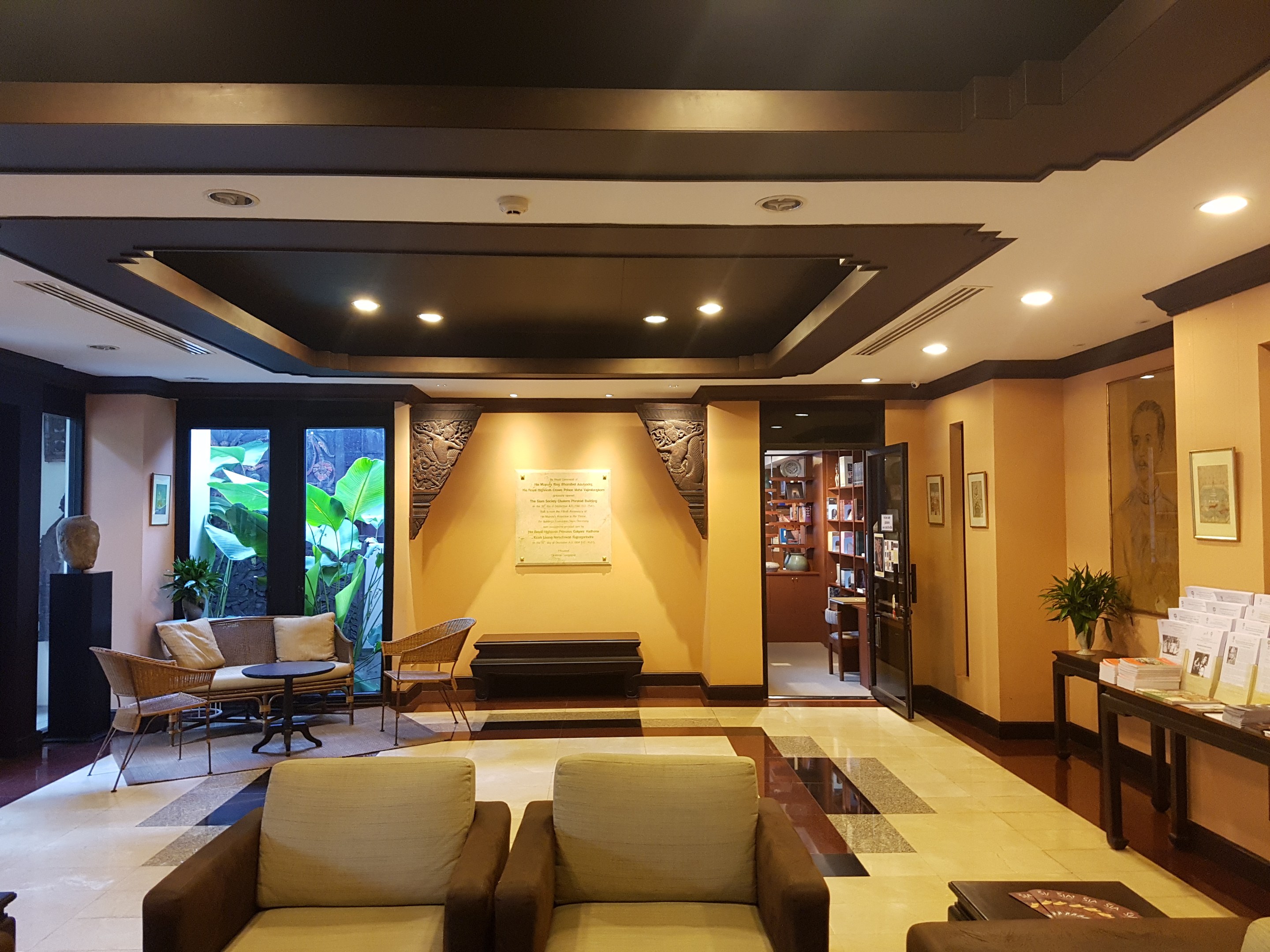 Ein geräumiger Hotelfoyer mit Sitzbereichen, Dekorationspflanzen, Bücherregalen, Leuchten, Wandkunst und Seitentisch-Dekoration, beleuchtet durch natürliches Licht von Fenstern und Türen.