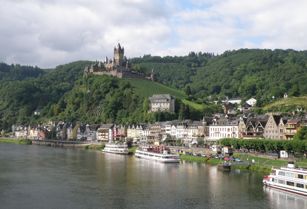 Ein malerischer Blick auf den Rhein in Deutschland mit einer Burg auf einem Hügel, Booten auf dem Fluss, Fahrzeugen auf einer näheren Straße und einem bewölktem Himmel.