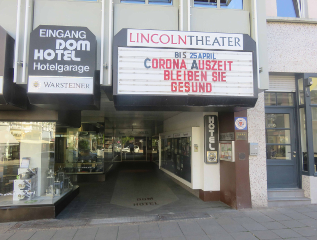 Außenansicht des Lincoln Theaters in Berlin, Deutschland, mit Glasfenstern und -türen sowie einer Tafel mit Text und einem belebten Stadtbild im Inneren.
