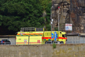 Ambulanz neben einer Straße an einem Abhang geparkt, mit einer Wand, Geländer, Pfählen, Lampen, Schildern und anderen Gegenständen im Vordergrund und Bäumen und einem Auto im Hintergrund.
