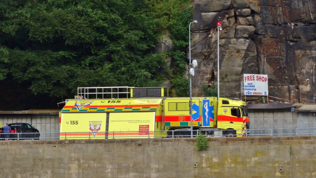 Ambulanz neben einer Straße an einem Abhang geparkt, mit einer Wand, Geländer, Pfählen, Lampen, Schildern und anderen Gegenständen im Vordergrund und Bäumen und einem Auto im Hintergrund.