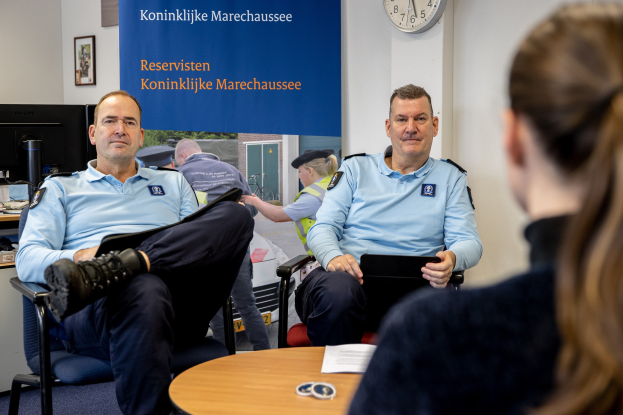 Zwei Polizeibeamte sitzen an einem Tisch und unterhalten sich mit einer Frau; im Hintergrund sind eine Wandtafel, eine Uhr und Foto Rahmen sowie ein weiterer Tisch mit einem Monitor, Tastatur und Maus zu sehen.