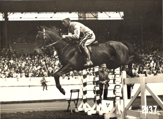 Ein Mann in einem weißen Kleid und Hut reitet auf einem braunen Pferd, springt über ein Hindernis, während Zuschauer aus dem Stadion zusehen.