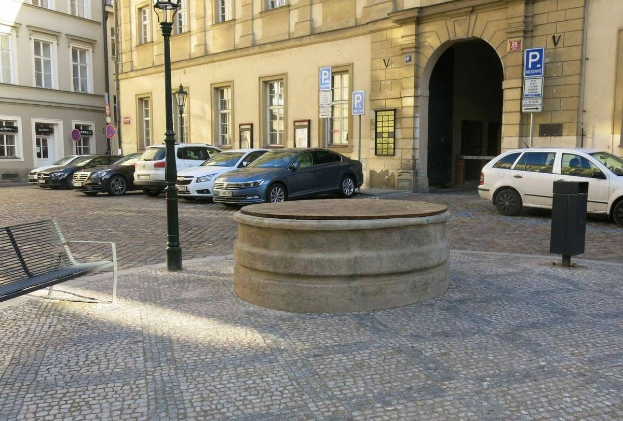Ein Stadtplatz mit einer zentralen Bank, umgeben von Gebäuden mit Fenstern, Laternenmasten und Schildern, und einem Springbrunnen in der Mitte.