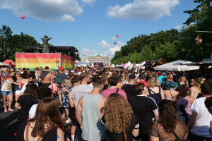 Eine große Menge geht eine Straße mit Zelten, Bäumen, Pfählen, Lichtern und einer Statue entlang während des Christopher Street Day Festivals in Berlin, mit Gebäuden, Wolken und Ballons im Hintergrund.
