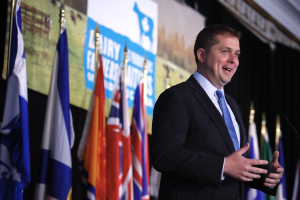 Ein Mann im Anzug und Krawatte spricht an einem Rednerpult mit Mikrofon, umgeben von Fahnen und einem Bildschirm, wahrscheinlich zu einer Regierungszahlung an die Milchindustrie.