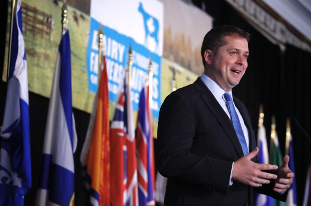 Ein Mann im Anzug und Krawatte spricht an einem Rednerpult mit Mikrofon, umgeben von Fahnen und einem Bildschirm, wahrscheinlich zu einer Regierungszahlung an die Milchindustrie.