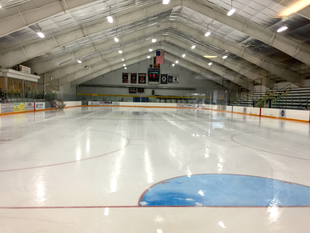 Große Indoor-Eisbahn mit Deckenbeleuchtung, Werbetafeln, einer Wand, Treppen mit Geländern und einigen Menschen, was auf eine Sportanlage hinweist, die likely für Hockey verwendet wird.