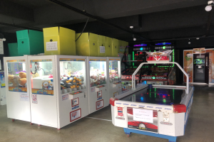 Ein Raum voller verschiedener Arcade-Spiele, mit einem Spielautomaten auf der rechten Seite und sichtbarem Boden sowie Deckenlampen darüber.