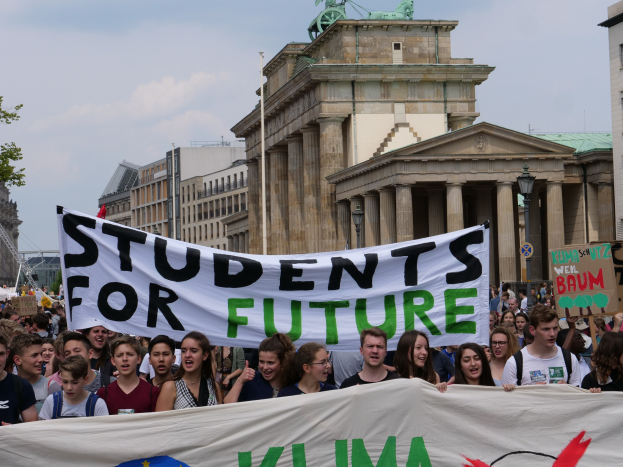 Eine Gruppe von Schülern marschiert in Berlin mit einer leuchtend bunten "Students for Future"-Schlagzeile vor dem Hintergrund von Gebäuden, Bäumen und Himmel.