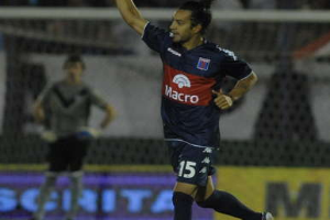 Ein Fussballspieler feiert auf dem Boden nach einem Tor, mit einer jubelnden Menge und einer Person im Hintergrund, alles vor einem leicht unscharfen Hintergrund.