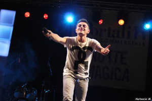 Ein Mann auf einer hell erleuchteten Bühne hält ein Mikrofon in der Hand, während im Hintergrund Musikinstrumente und ein "Nouveau Magica"-Schild zu sehen sind.