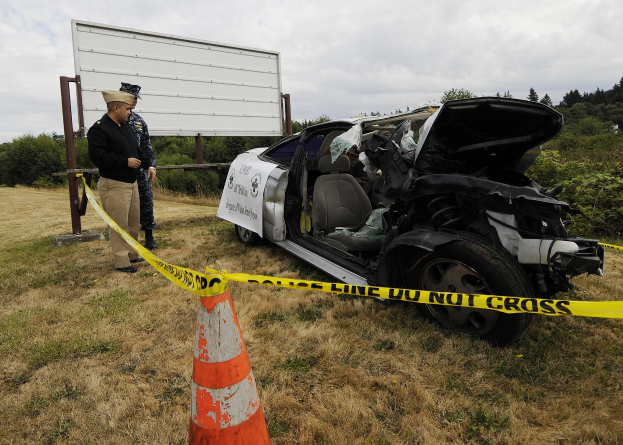 Ein Polizist in Uniform steht neben einem schwer beschädigten Auto auf einem grasbewachsenen Straßenrand, mit eingedellter Motorhaube, zersplitterten Fenstern und einem Verkehrskegel und Band in der Nähe gegen den Hintergrund von Bäumen und einem bewölkten Himmel.