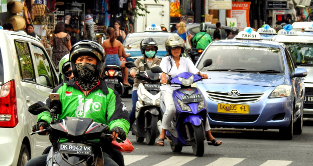 Eine Gruppe von Menschen auf Motorrädern fährt eine belebte Straße entlang mit Autos, Gebäuden, Bäumen und Geschäften im Hintergrund.