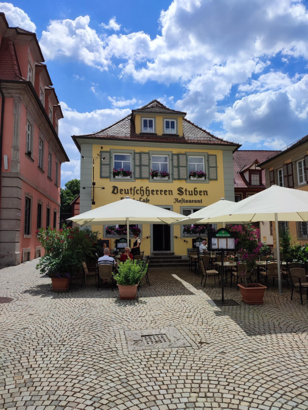 Freisitz mit Tischen und Sonnenschirmen vor einem gelben Gebäude mit der Aufschrift "Deutschen Stuben Restaurant" in Baden-Württemberg, Deutschland, mit einigen Menschen, die sitzen und stehen, unter einem blauen und weißen Himmel.