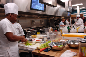 Eine Gruppe von Köchen in einer Küche beim Zubereiten von Speisen auf einem Schneidbrett, mit einem Koch im Vordergrund in einer weißen Schürze und Mütze, der ein Messer hält und von Utensilien, Schüsseln, Gemüse, einem Handy, Flaschen und anderen Gegenständen umgeben ist, mit einem Fernseher und Deckenlampen im Hintergrund.