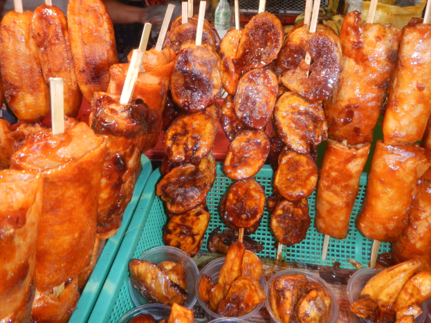 Ein Marktstand mit Spießen von Hähnchenflügeln und anderen Lebensmitteln in Plastikbehältern, mit Menschen und Flaschen im Hintergrund.