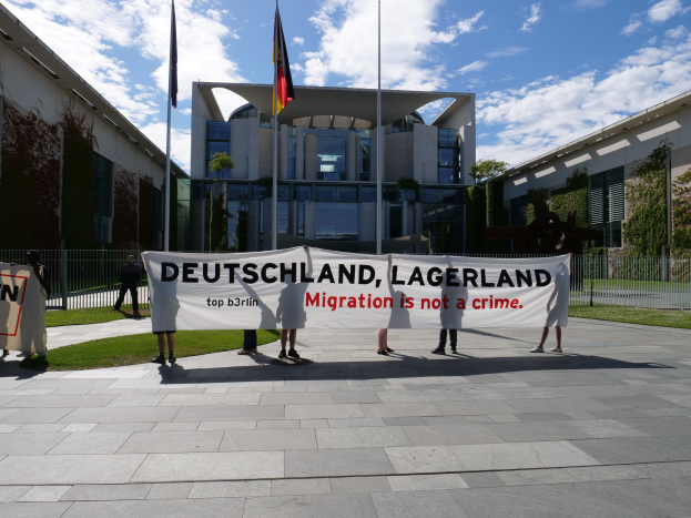 Gruppe von Menschen mit einem Transparent "Deutschland, Lagerland Migration ist kein Verbrechen" vor einem Gebäude mit Fenstern, umgeben von Gras, einem Metallzaun, Pflanzen, Bäumen und Fahnenstangen unter einem bewölkten Himmel.