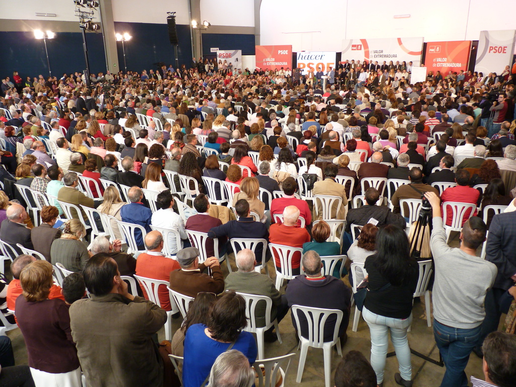Ein volles Stadion mit vielen sitzenden und stehenden Menschen, eine Bühne mit zwei Rednern an einem Podium und verschiedene Banner und Lichter.