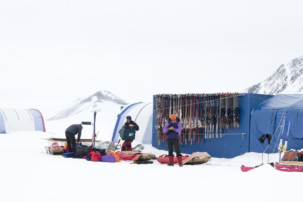 Drei Personen stehen auf einer schneebedeckten Landschaft mit verstreuten Taschen, Zelten mit Skiern dahinter und schneebedeckten Hügeln im Hintergrund unter einem klaren Himmel.