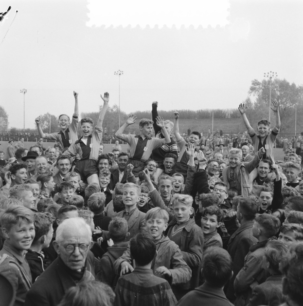 Schwarzes und weißes Foto einer Menge vor einem Stadion, einige halten Hände zum Jubeln hoch, mit Laternenmasten, Bäumen und einem klaren Himmel im Hintergrund.