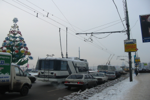 Eine belebte Stadtstraße mit Fahrzeugen, einem geschmückten Weihnachtsbaum, Schnee auf dem Boden, Fußgängern und Gebäuden unter einem klaren Himmel.