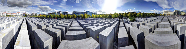 Luftaufnahme des Holocaust-Mahnmals für die ermordeten Juden Europas in Berlin, Deutschland, mit einem Gitter aus Betonsteinen, umgeben von Bäumen, Gebäuden und Fahrzeugen auf einer Straße mit einem bewölkten Himmel.