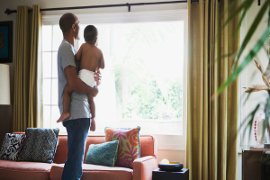 Ein Mann trägt ein Kind vor einem Fenster mit Vorhängen, einem Sofa und mehreren Kissen.
