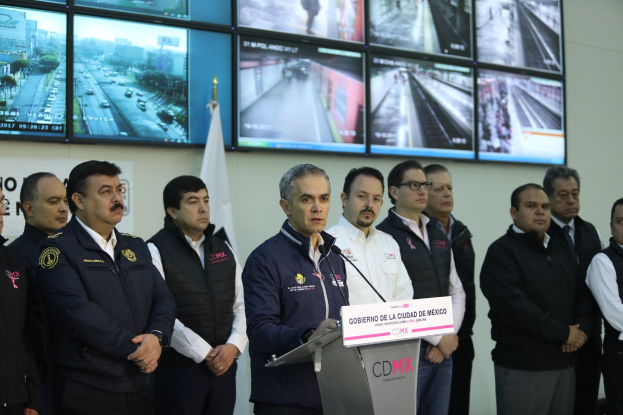 Eine Gruppe von Männern, die vor einem Rednerpult mit einem Mikrofon und einer Tafel mit der Aufschrift 'Mexikanische Zoll- und Grenzschutzbehörde' stehen, mit Fernsehgeräten und einer Flagge an der Wand dahinter.