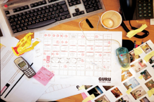 Ein unordentlicher Schreibtisch mit Papieren, einem Computer, einem Telefon, Tassen und einem Handy.