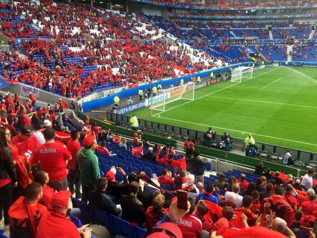 Eine große Menge in einem hell erleuchteten Stadion schaut einem Fußballspiel zu, mit Menschen, die sitzen und stehen, Schildern und einem Torpfosten im Hintergrund.