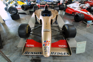 Rennwagen in einem Motorsportmuseum ausgestellt, getrennt durch einen Metallzaun mit Informationsschild im Hintergrund.