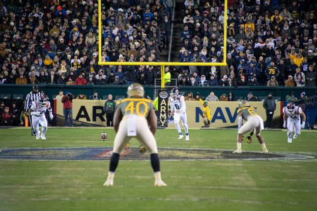 Fußballspieler mit Helmen und Uniformen auf dem Feld mit Zuschauern auf den Tribünen und Stadionwerbung im Hintergrund.