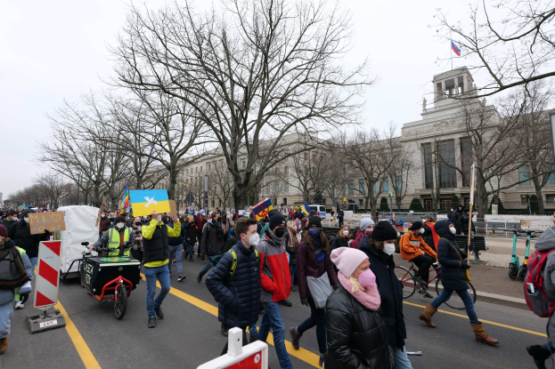 Eine große Gruppe von Menschen marschiert auf einer Demonstration die Straße in Washington, D.C. entlang, einige halten Schilder und Banner, andere fahren Fahrräder, mit Bäumen und einem klaren blauen Himmel im Hintergrund.