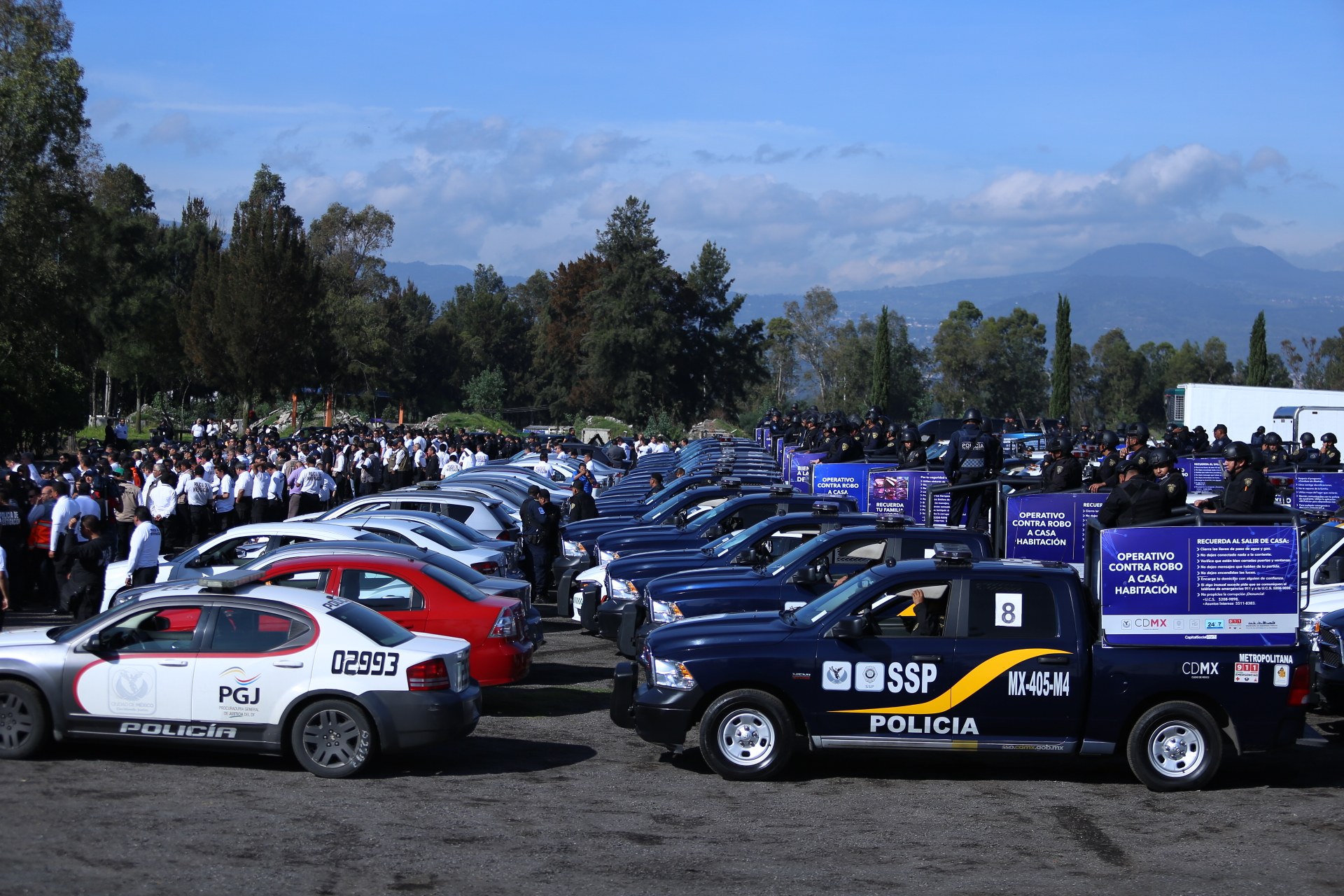 Eine große Anzahl von Polizeiwagen auf einem Parkplatz mit Menschen in der Nähe, Schilder mit Text und ein Hintergrund aus Bäumen, Hügeln und einer bewölkten Himmel.