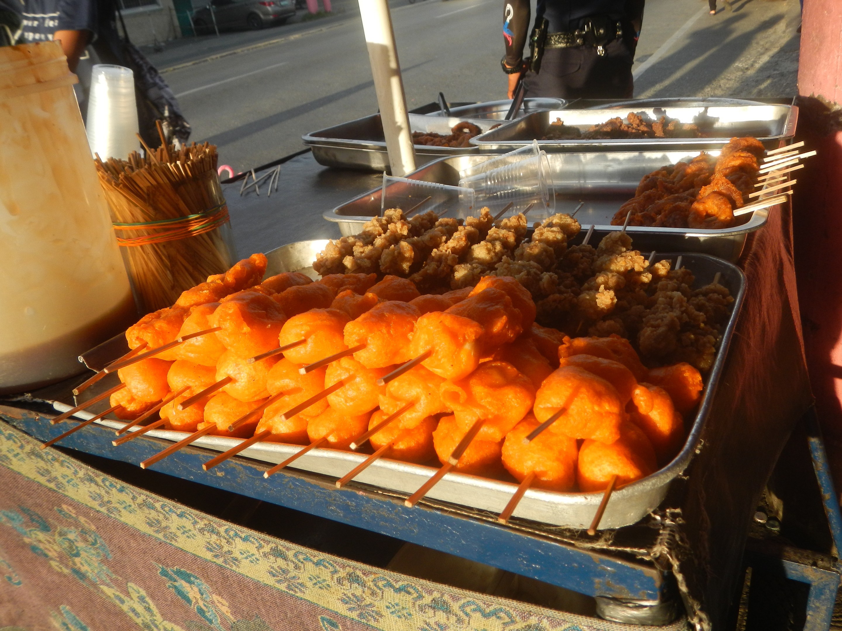 Ein Straßenhändler zeigt Tabletts mit Lebensmitteln auf einem Tisch mit Zahnstochern, einem Eimer und anderen Gegenständen, mit Menschen, Fahrzeugen, Pfählen, Gebäuden, Bäumen und einem klaren blauen Himmel im Hintergrund.