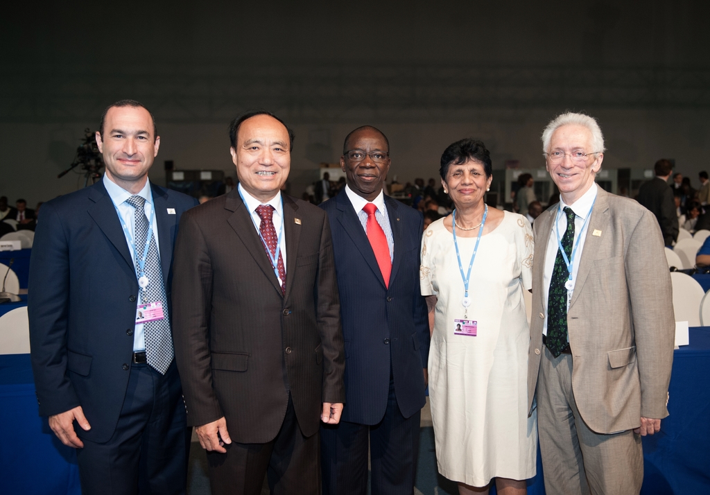 Eine Gruppe von Männern in Anzügen und eine Frau in einem Kleid stehen und lächeln in der Vordergrund eines Saals, mit Menschen, die auf Stühlen sitzen und stehen in der Mitte, Tischen und einem Mikrofon auf einem Tisch links und einer Wand mit anderen Gegenständen im Hintergrund.