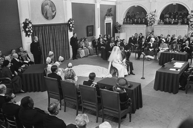 Ein Schwarz-Weiß-Foto einer Hochzeitszeremonie in einem großen Raum mit Gästen auf Stühlen und Stehplätzen, Tischen mit Papieren und Mikrofonen, einer dekorierten Wand mit Rahmen und Blumenbouquets sowie einem Podium mit Mikrofonständer.