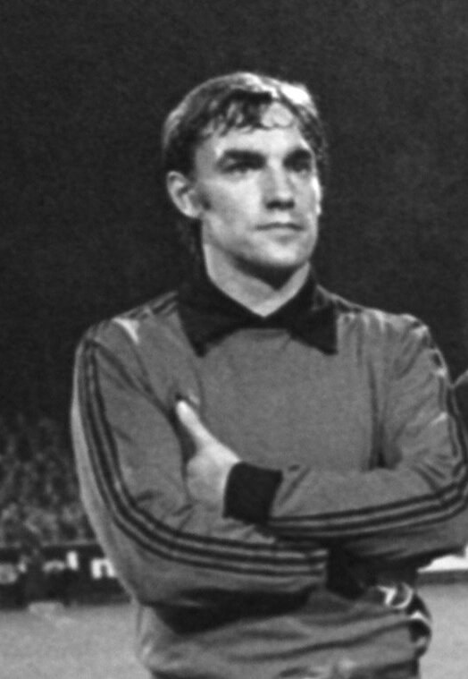 Schwarzes und weißes Foto eines Mannes auf einem Fußballfeld mit verschränkten Armen, umgeben von einer Gruppe sitzender Menschen im Hintergrund.