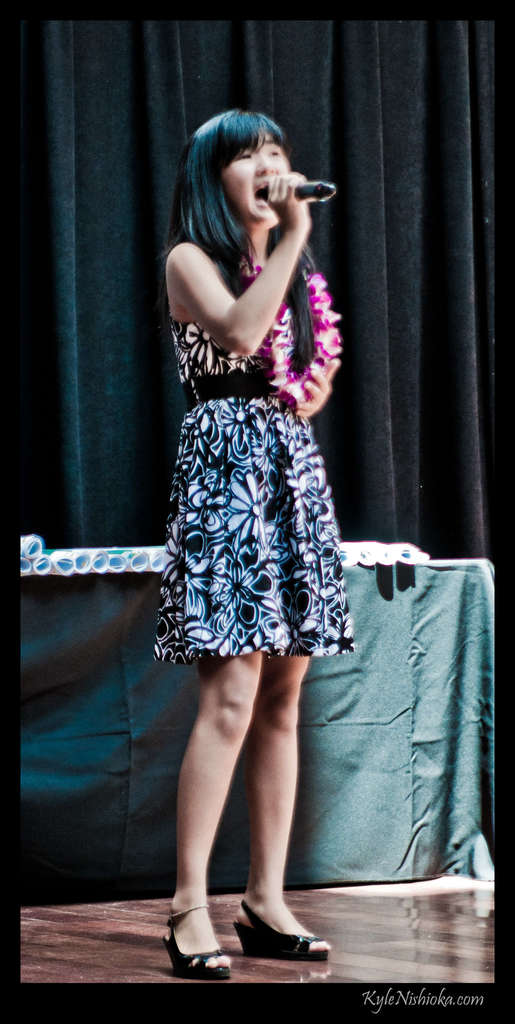 Eine Frau in einem bunten Kleid singt in ein Mikrofon auf der Bühne, mit einem Tisch voller Papiere und einem Vorhang im Hintergrund.