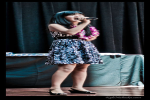 Eine Frau in einem bunten Kleid singt in ein Mikrofon auf der Bühne, mit einem Tisch voller Papiere und einem Vorhang im Hintergrund.